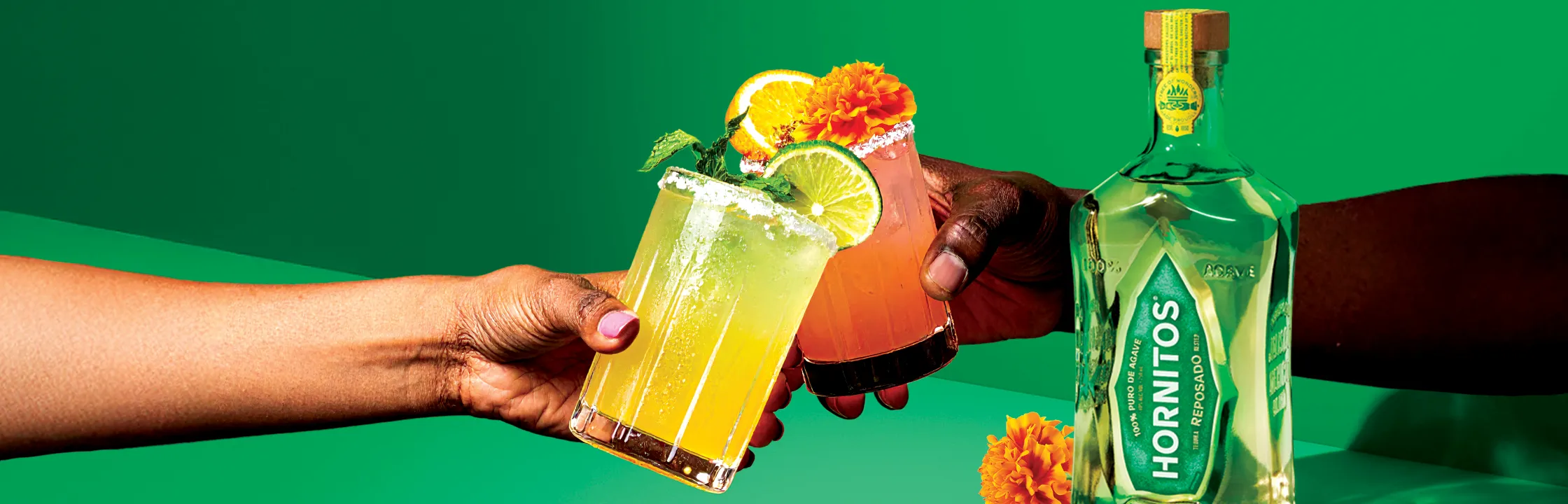 Two hands holding cocktails with lime and orange garnishes, against a green backdrop. A bottle of Hornitos tequila is positioned to the right.